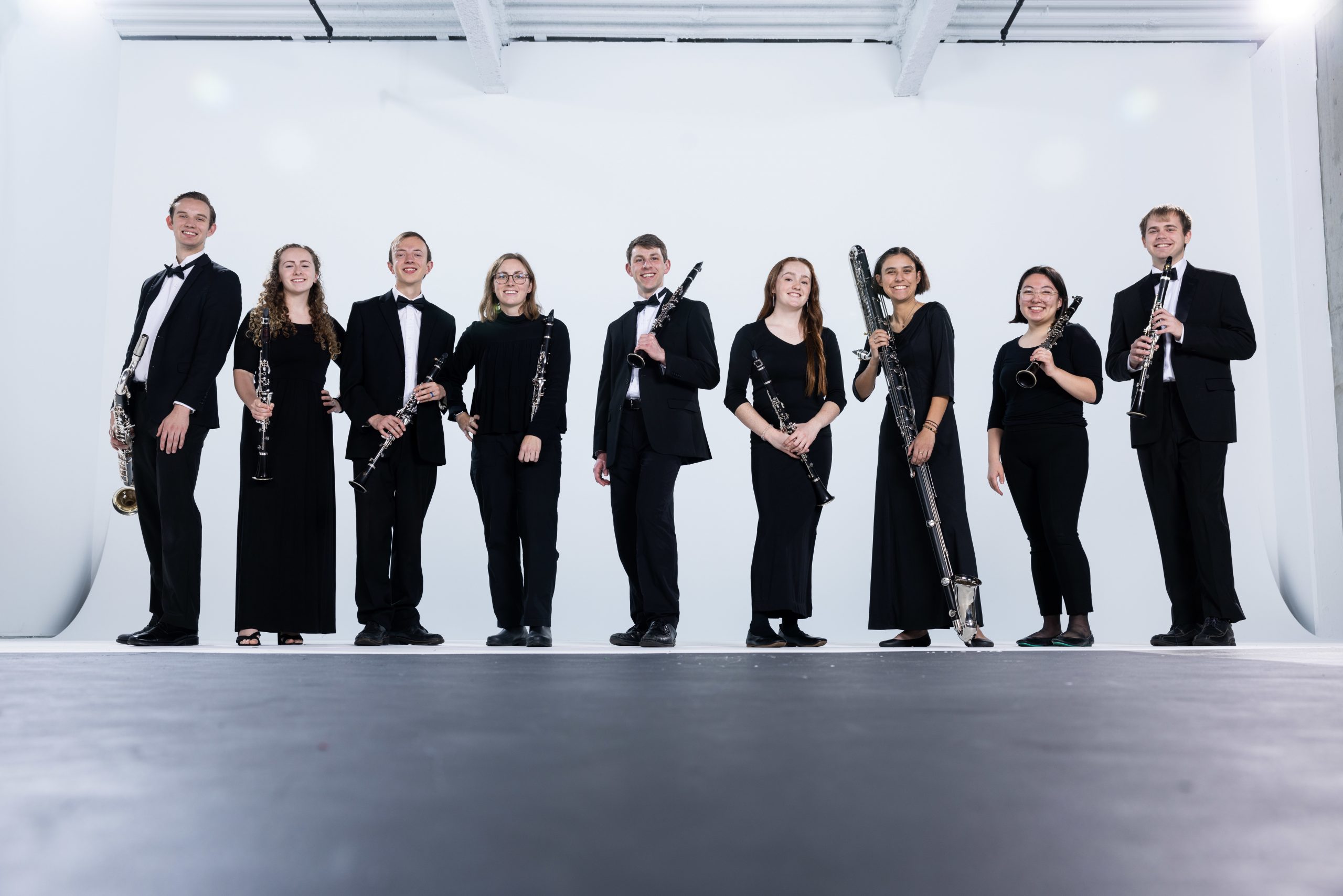 BYU Wind Symphony members stand in a line in front of a white, studio backdrop. The group smiles with their instruments while wearing concert black.