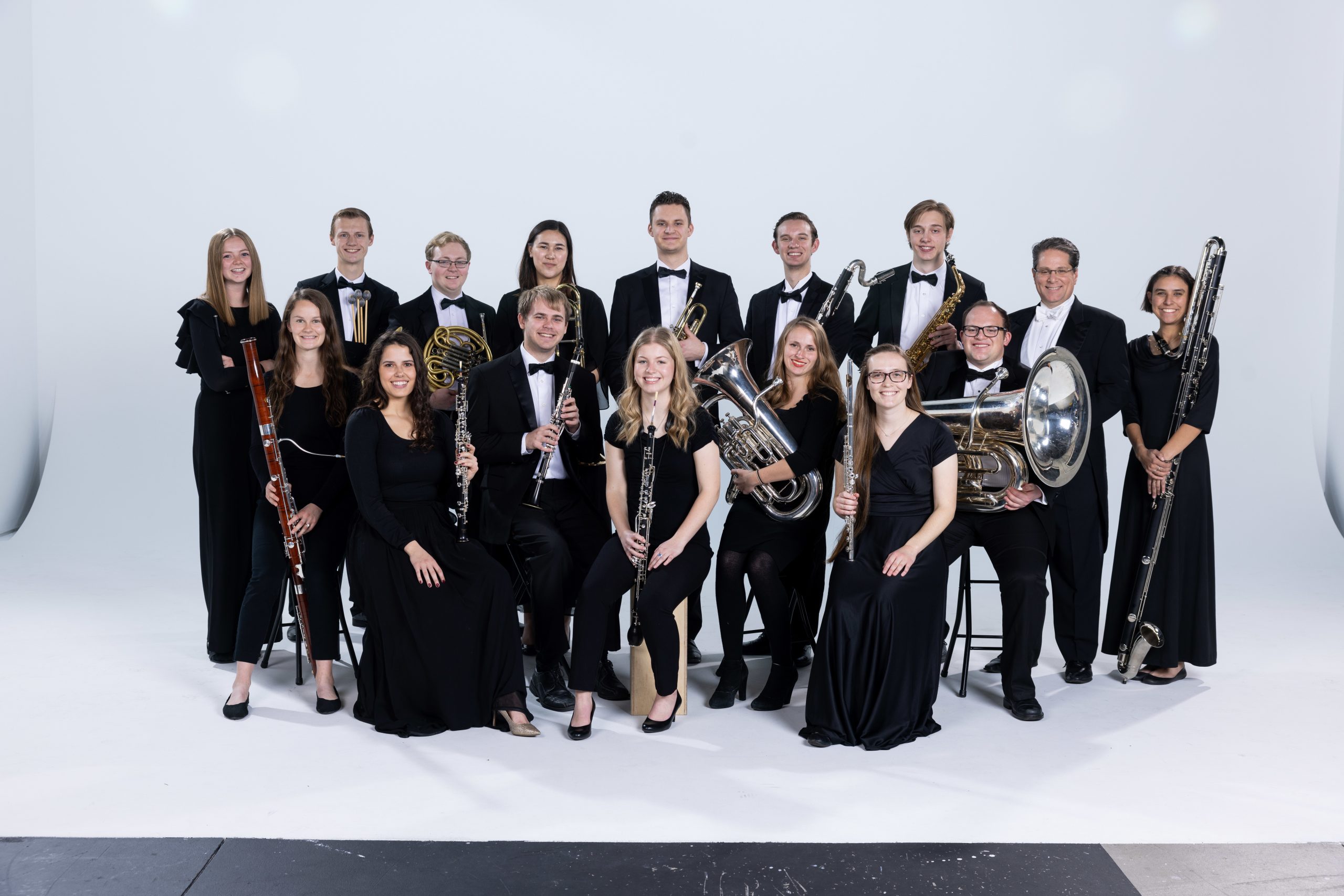 BYU Wind Symphony members gather around in concert black against a white studio back drop. The front row of musicians sit with instruments in their laps.