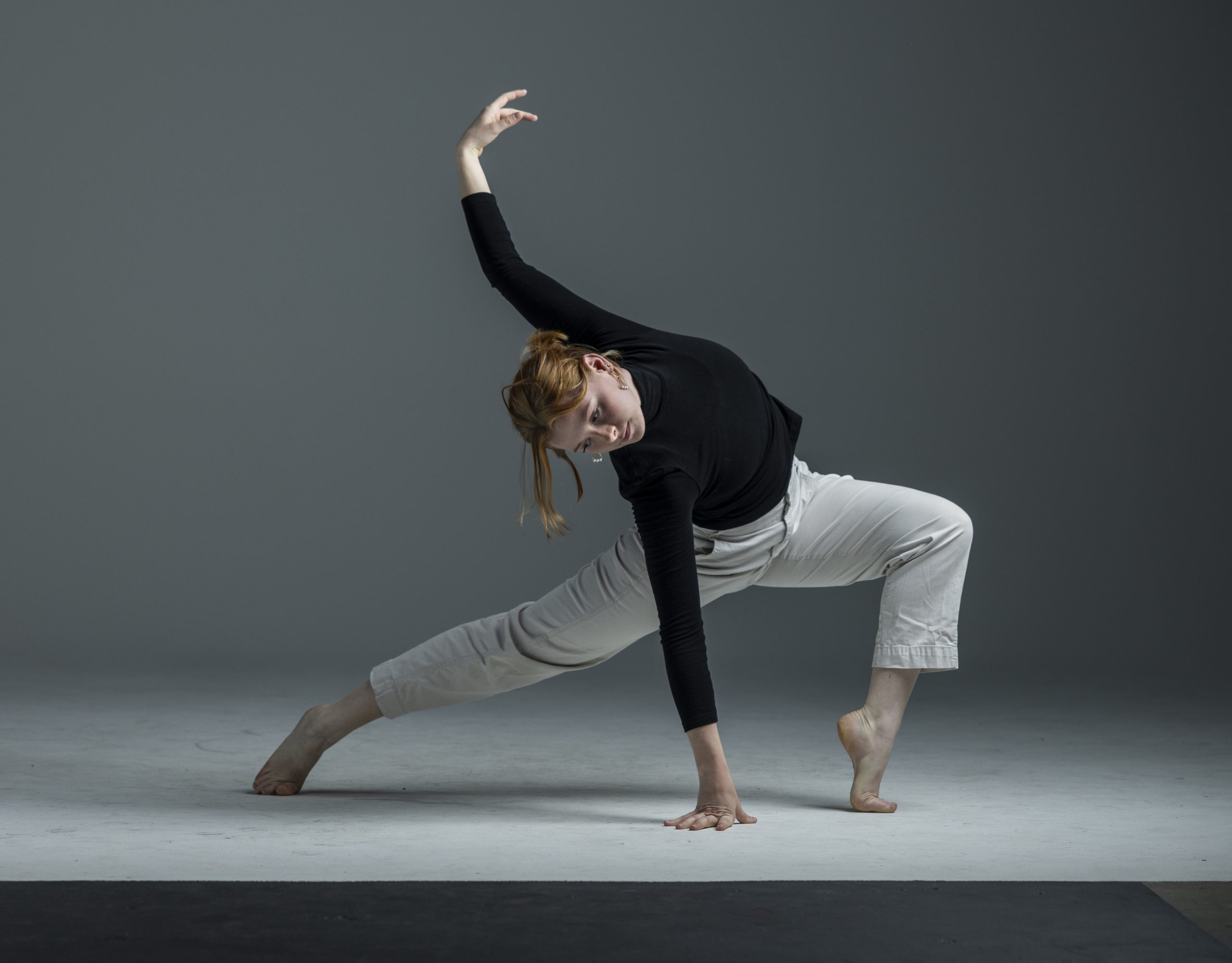 A contemporary dancer performs in a studio, balancing low to the ground. She extends one leg out to the side, supporting herself with one hand on the floor while the other arm arches overhead. She wears a black long-sleeve top and light pants, dancing barefoot against a gray background.