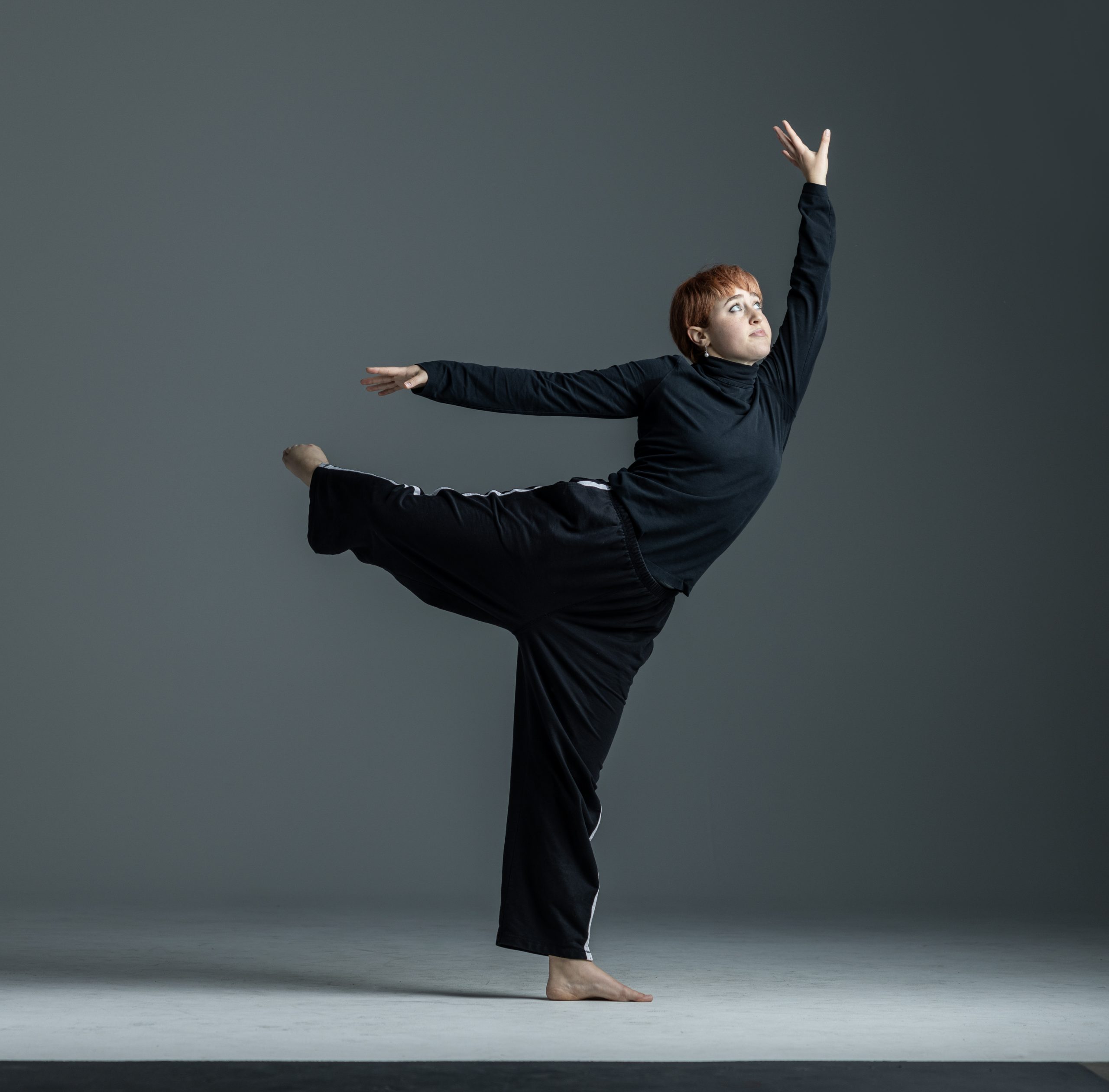 A contemporary dancer performs in a studio, balancing on one leg with the other extended straight back. One arm reaches forward while the other stretches upward, creating a dynamic diagonal line. The dancer wears a black long-sleeve top and loose black pants, moving barefoot against a gray background.