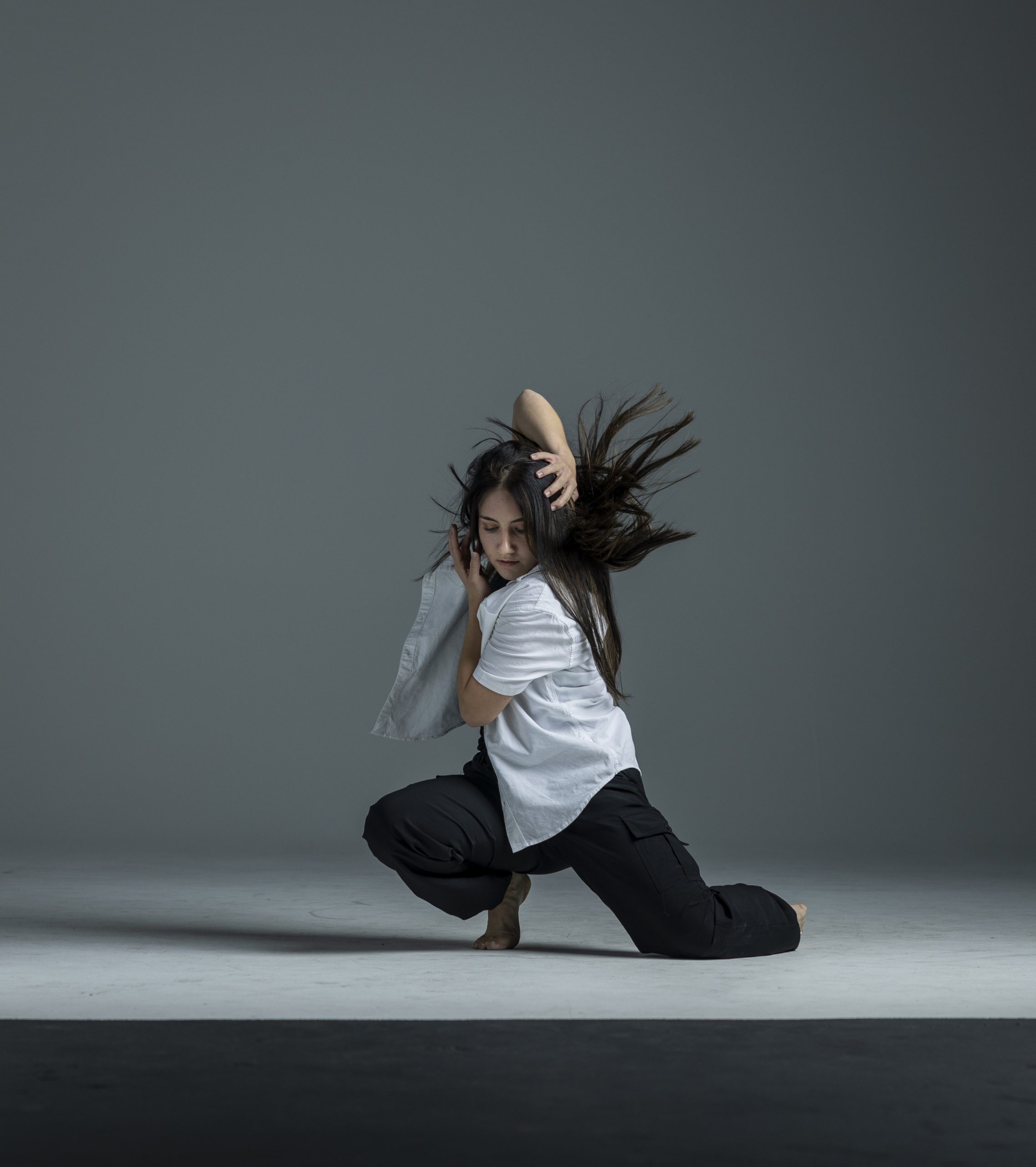 A contemporary dancer performs in a studio, kneeling low with one leg extended as her hair flies in motion. She curves her body inward, with one hand pressed to her head and the other framing her face. She wears a loose white shirt and black pants, dancing barefoot against a gray background.