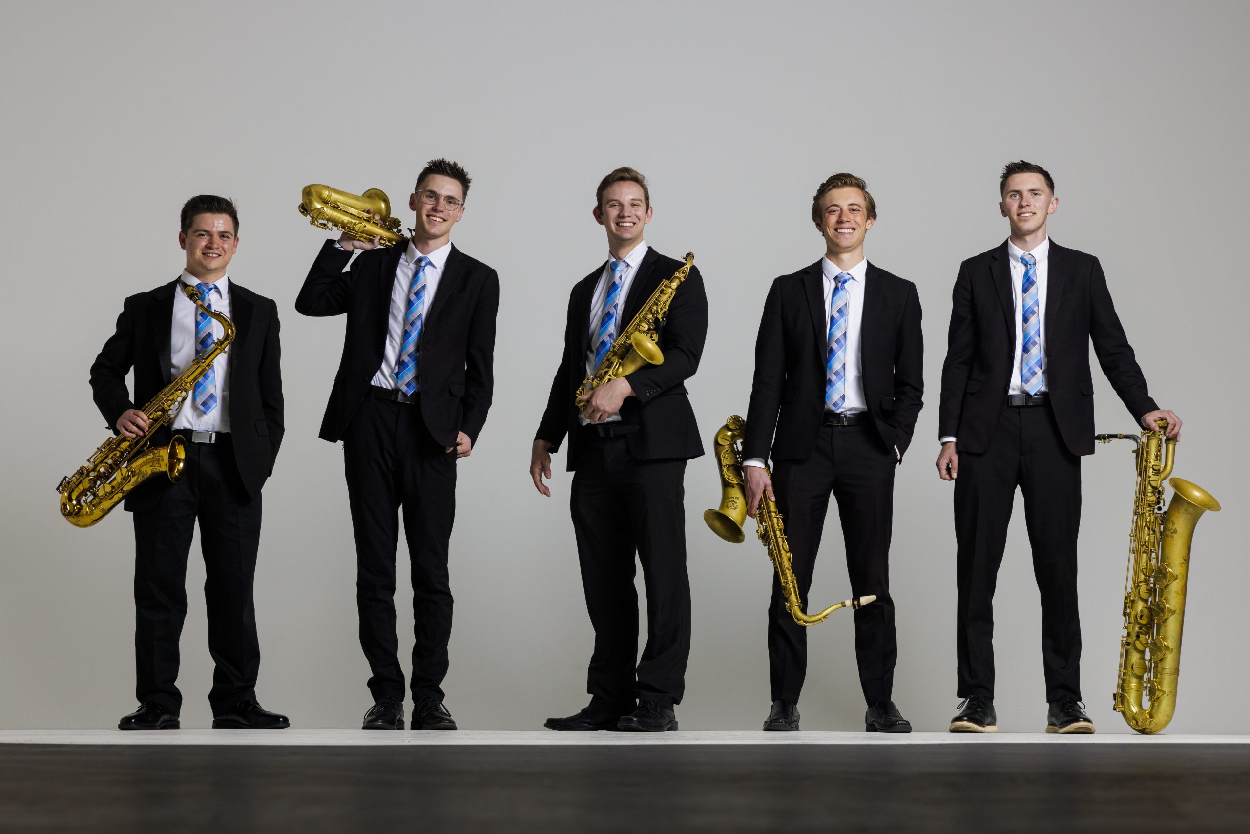 BYU Synthesis wind section poses in concert attire against a white, studio backdrop, smiling and holding their instruments