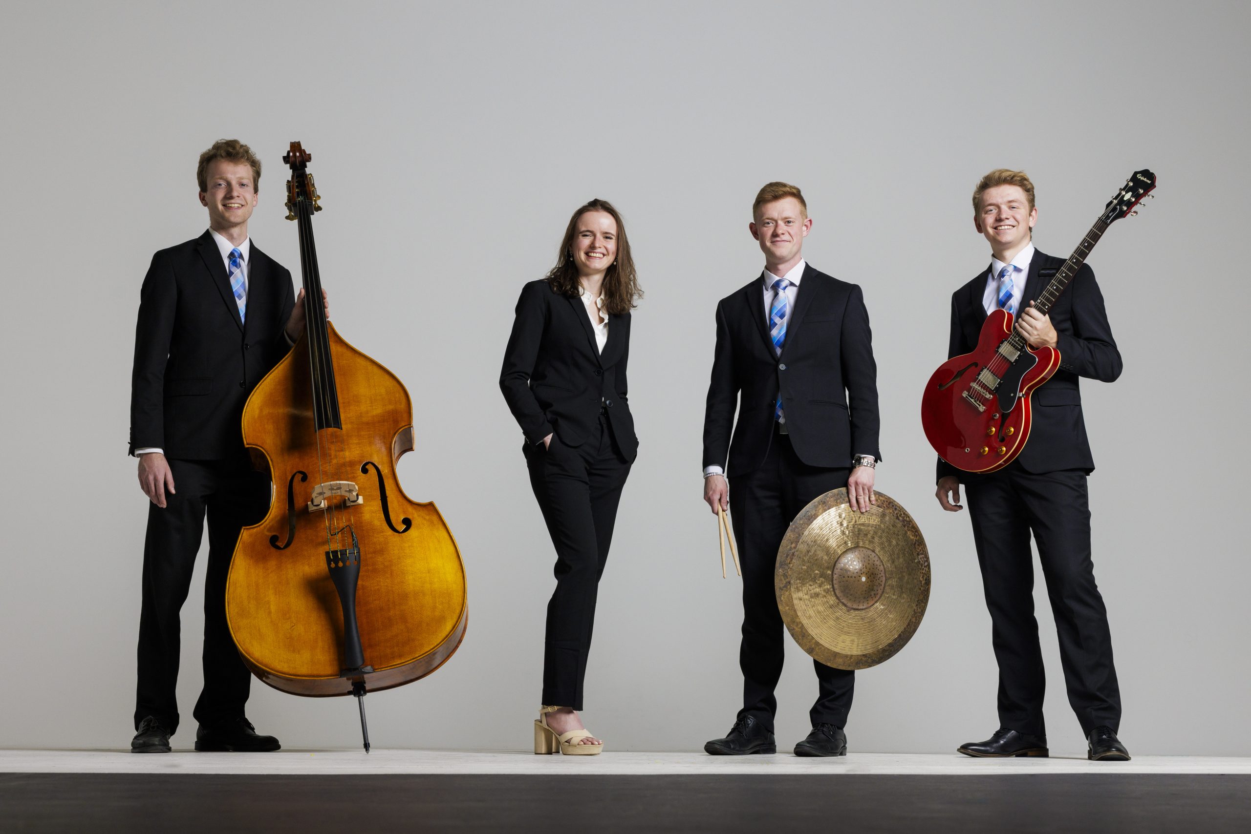 BYU Synthesis rhythm section stands in concert attire smiling, holding their instruments against a white studio backdrop.