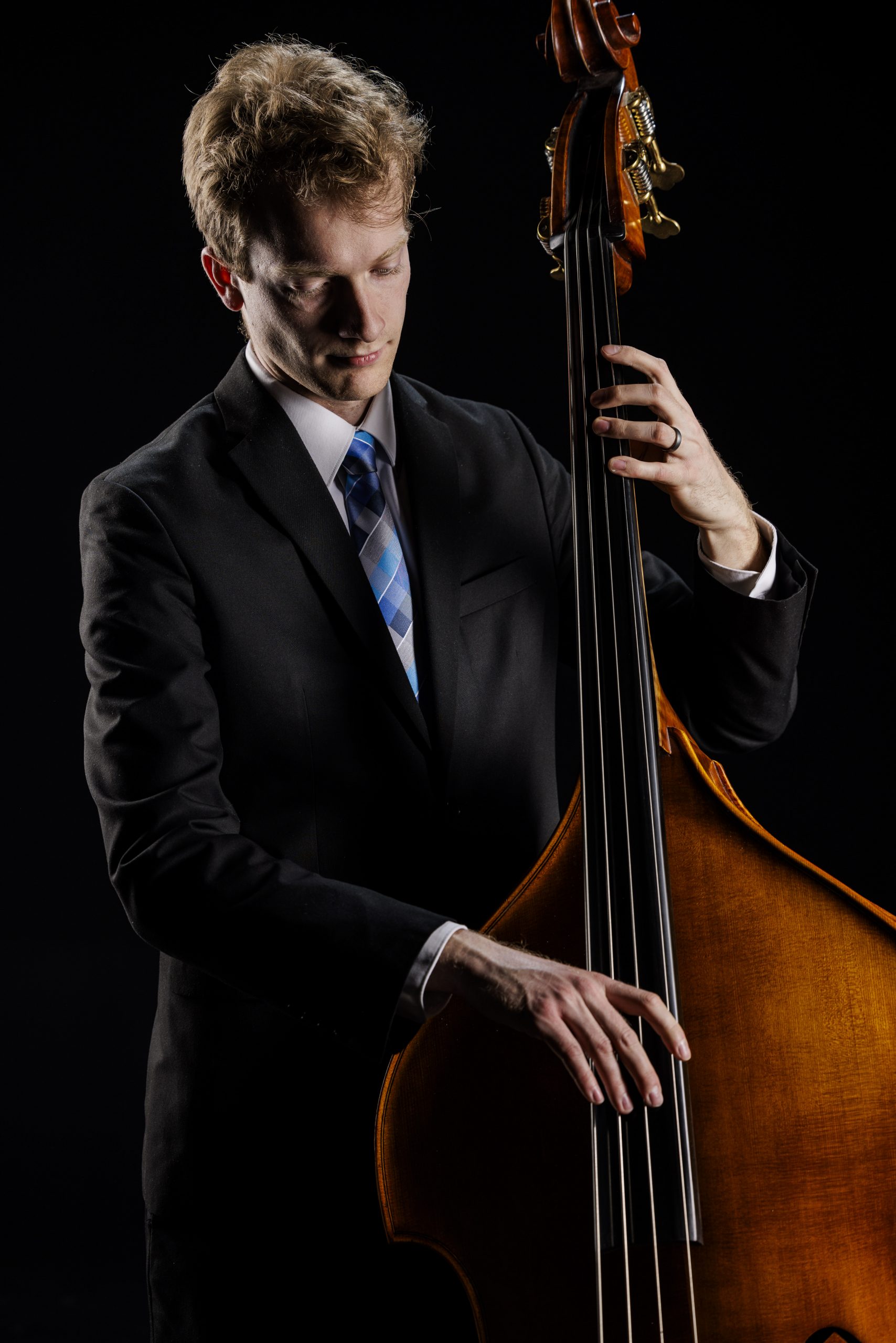 BYU Synthesis upright bass player plucks a string while standing against a black studio background wearing concert attire