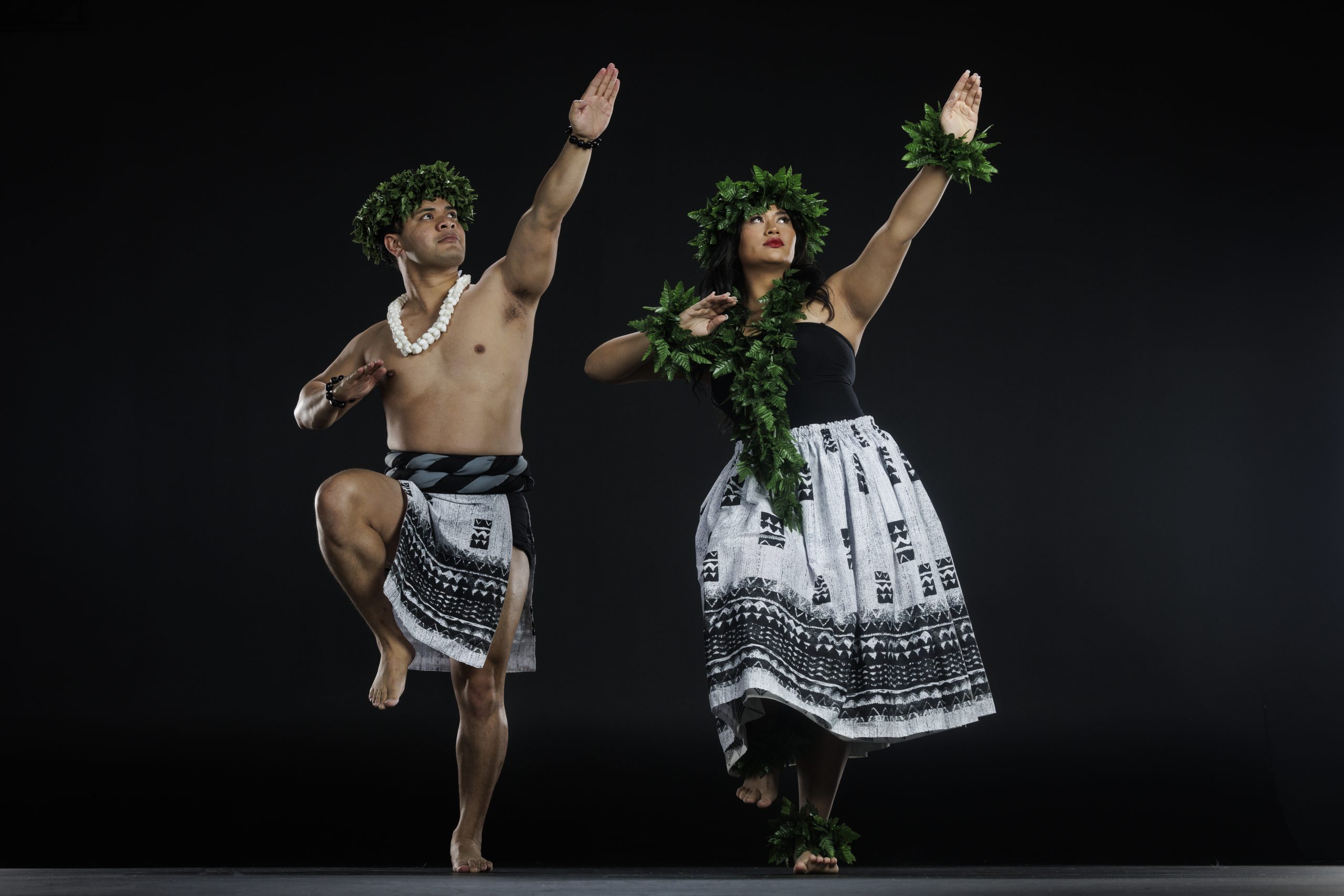 Two dancers in traditional Polynesian attire pose in a studio. The male dancer, wearing a leaf headpiece, shell necklace, and patterned skirt, stands on one leg with one arm extended upward. The female dancer, wearing a leafy crown, wrist, and ankle adornments with a black top and patterned skirt, mirrors the upward gesture. Both are barefoot against a plain dark background.