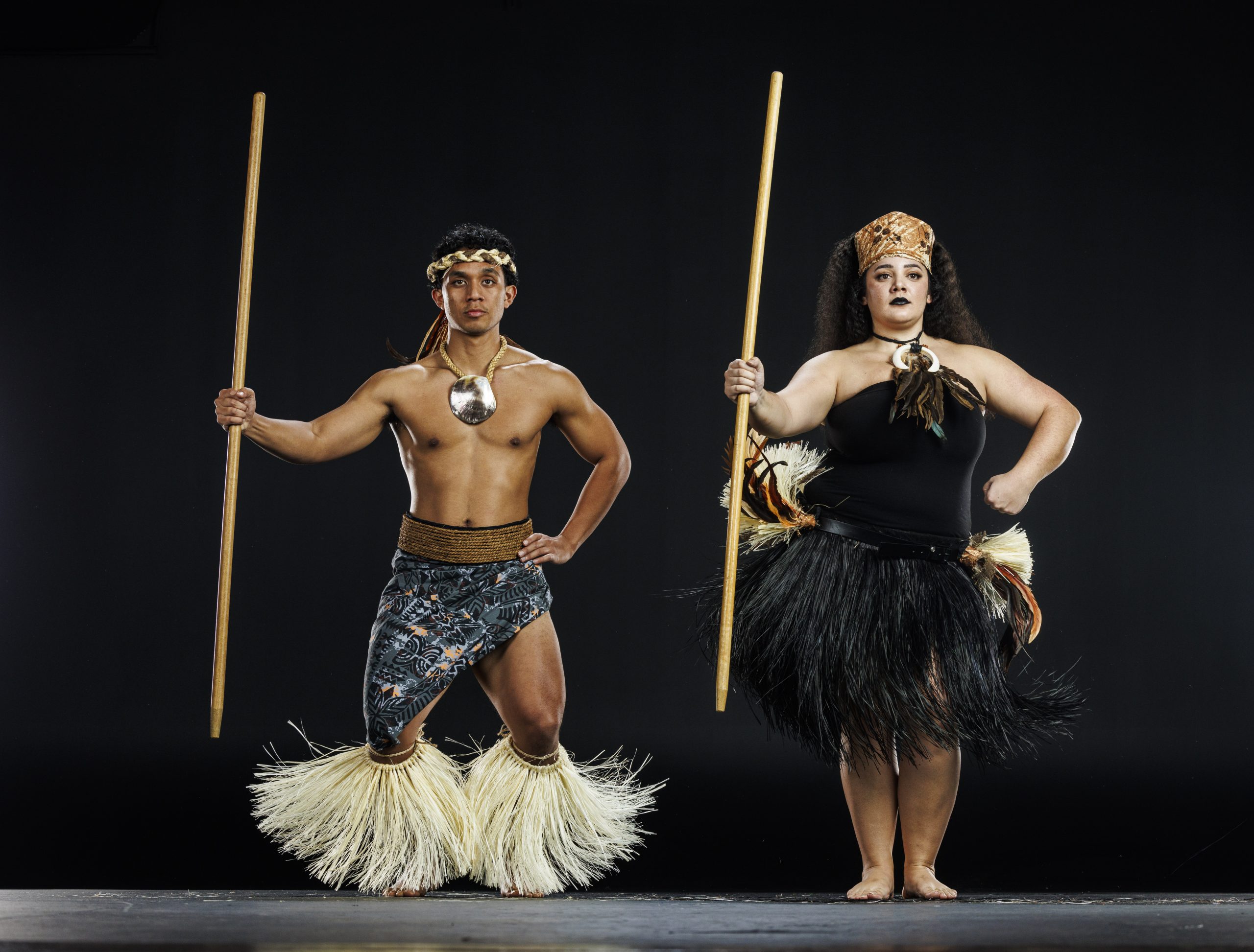 Two dancers in traditional Polynesian attire pose in a studio holding long wooden staffs. The male dancer, bare-chested with a shell necklace, patterned wrap, and straw leg adornments, squats with one hand on his hip. The female dancer wears a headdress, black top, feathered necklace, and a skirt made of black and natural fibers, standing firmly with one hand on her hip. Both are barefoot against a plain dark background.