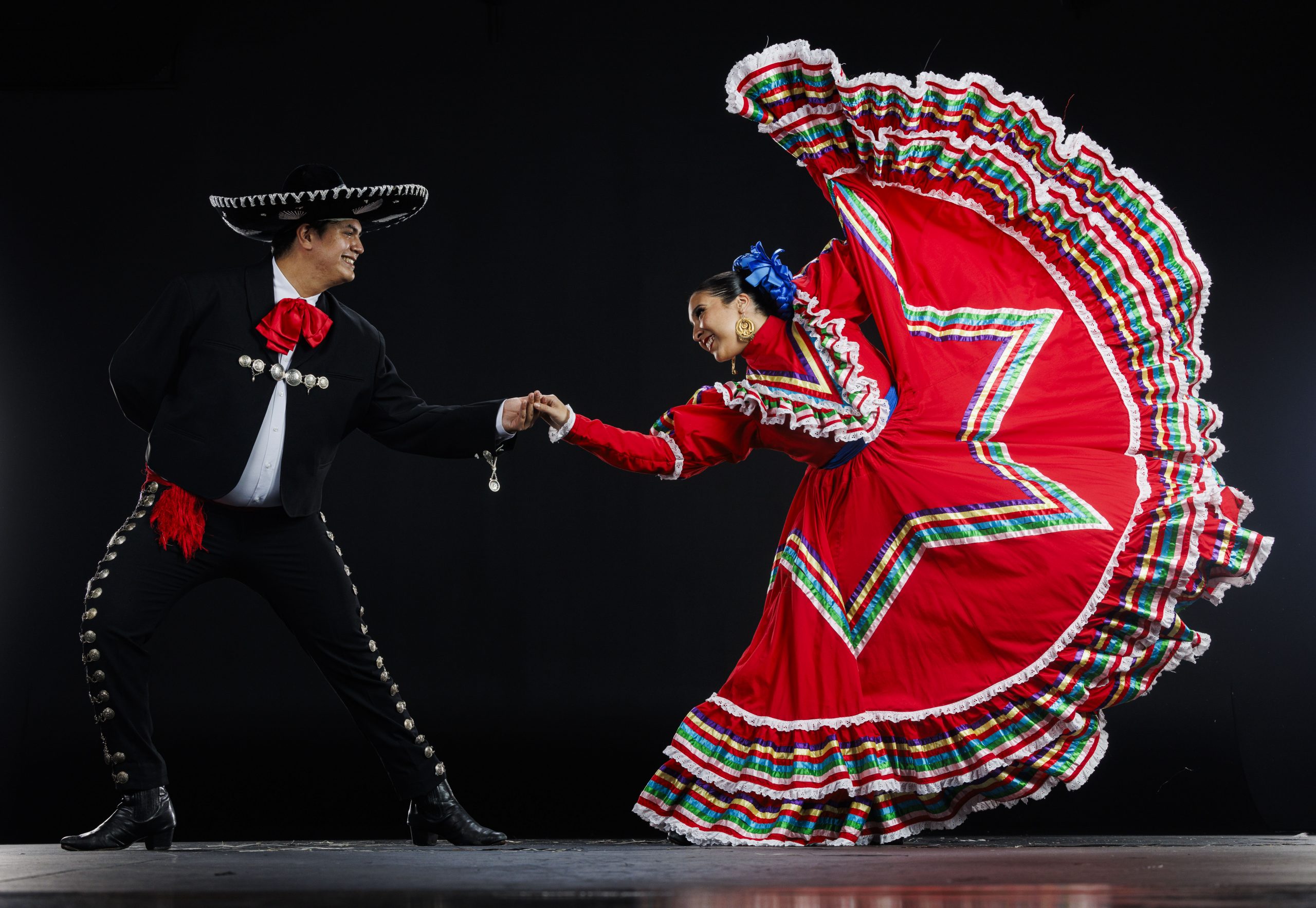 Two dancers in traditional Mexican folklórico attire pose in a studio. The male dancer wears a black charro suit with silver buttons, a red tie, and a wide-brimmed sombrero. The female dancer wears a bright red dress with multicolored ribbon accents and holds the skirt out in a dramatic flourish. They smile at each other while holding hands, standing against a plain dark background.