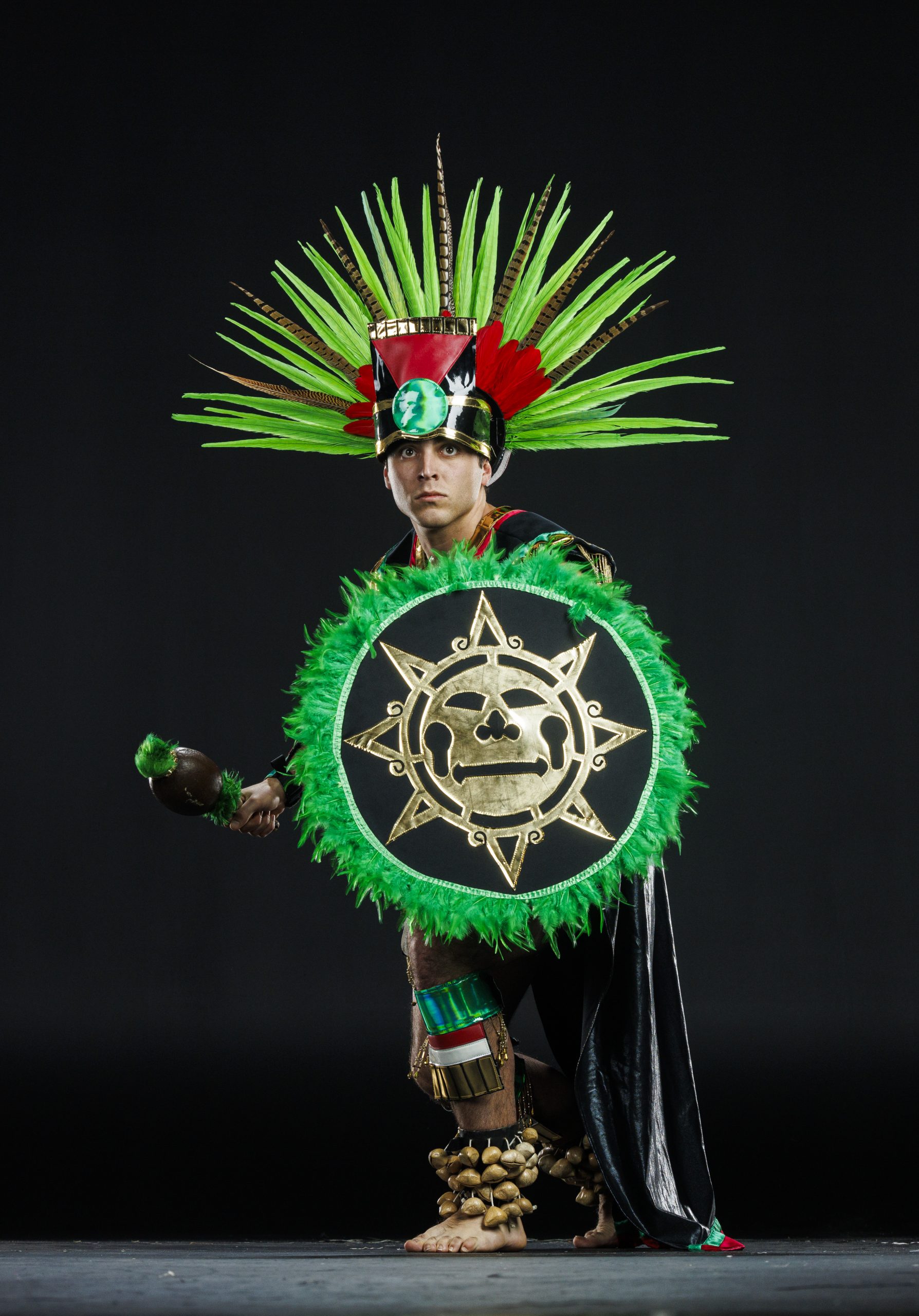 A dancer in Native American regalia poses in a studio. He wears a large feathered headdress with green, red, and brown plumes, a black cape, and ankle rattles. He holds a decorated green-and-gold shield with a sun design in one hand and a ceremonial rattle in the other. He stands barefoot against a plain dark background.