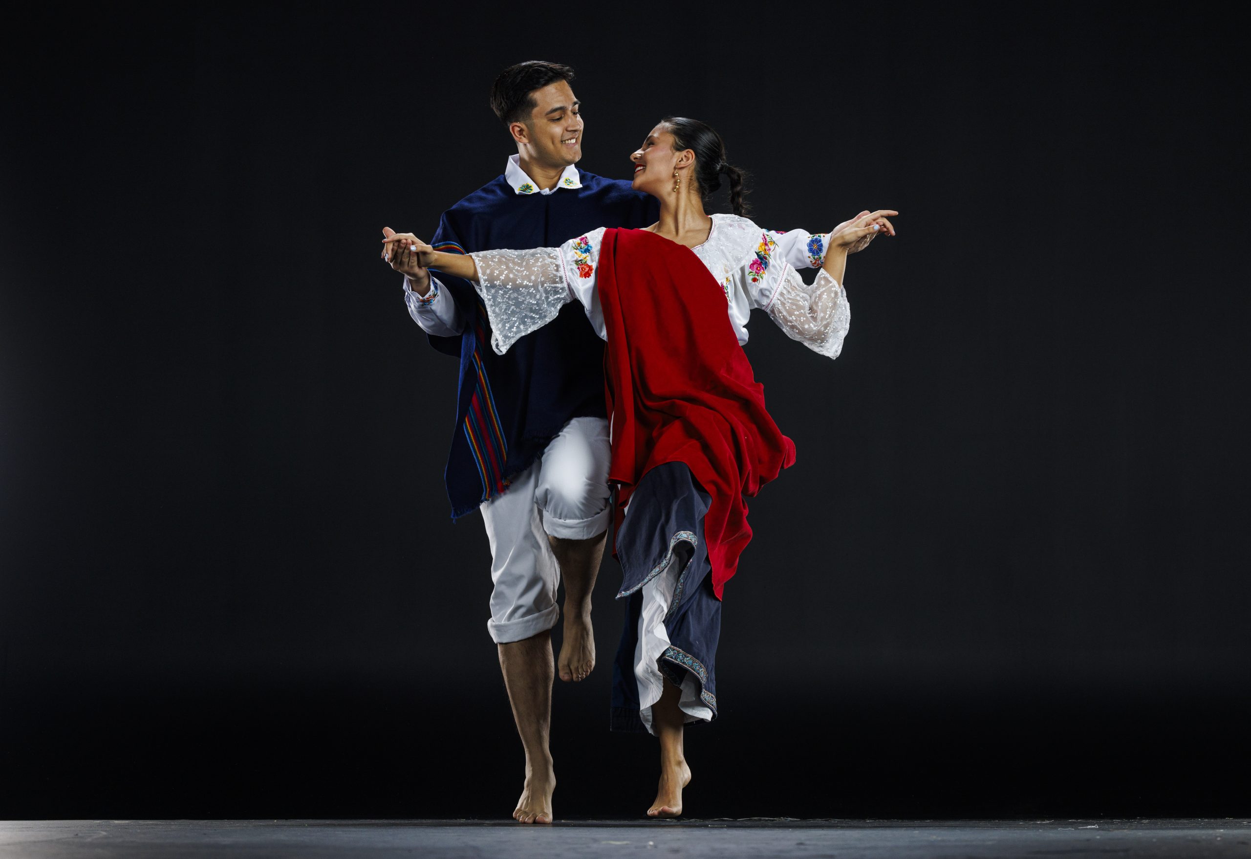 Two dancers in traditional Latin American attire perform in a studio. The male dancer wears a navy poncho with red stripes, a white shirt, and cropped white pants. The female dancer wears a white blouse with embroidered flowers, a red shawl, and a long skirt. They smile at each other while holding hands, dancing barefoot against a plain dark background.