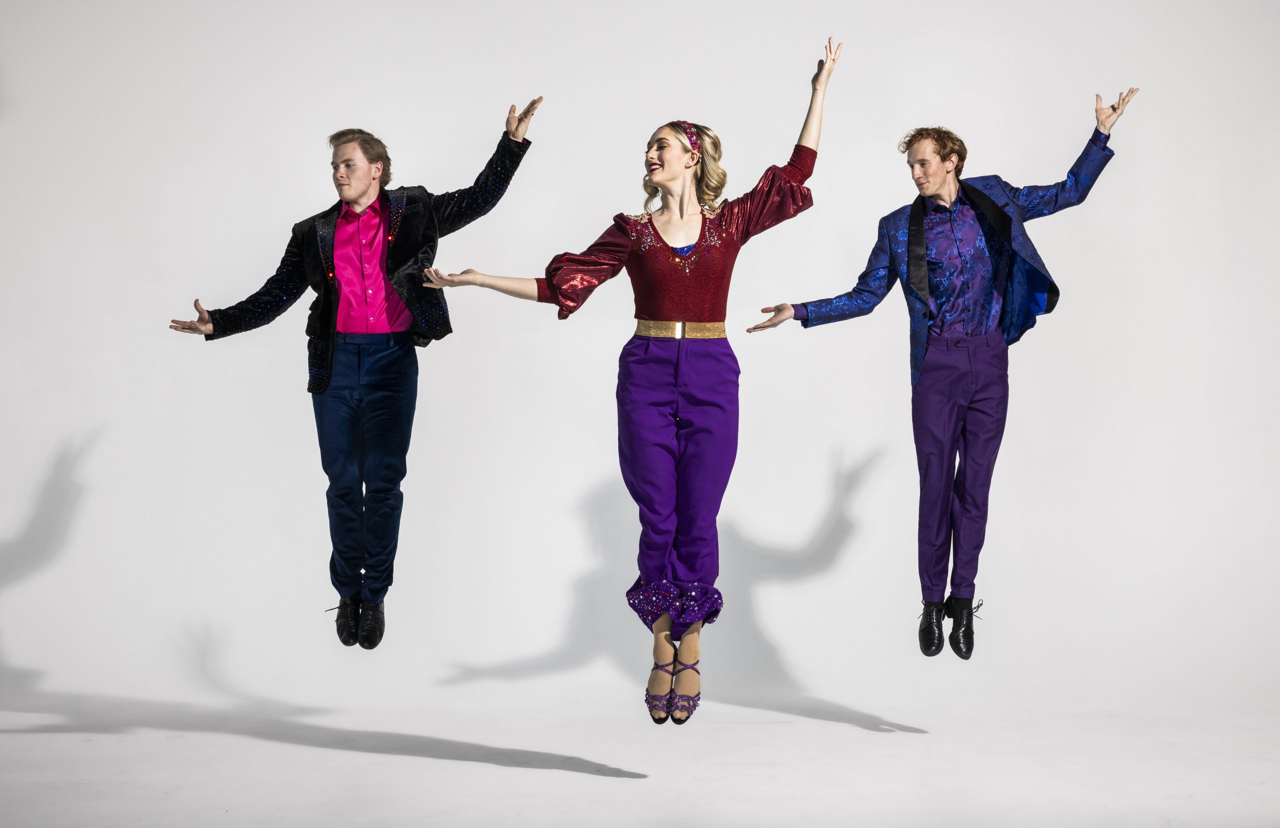 Three dancers strike the same pose midair, arms extended to create upward movement. They wear costumes featuring bold colors and flashy accents.