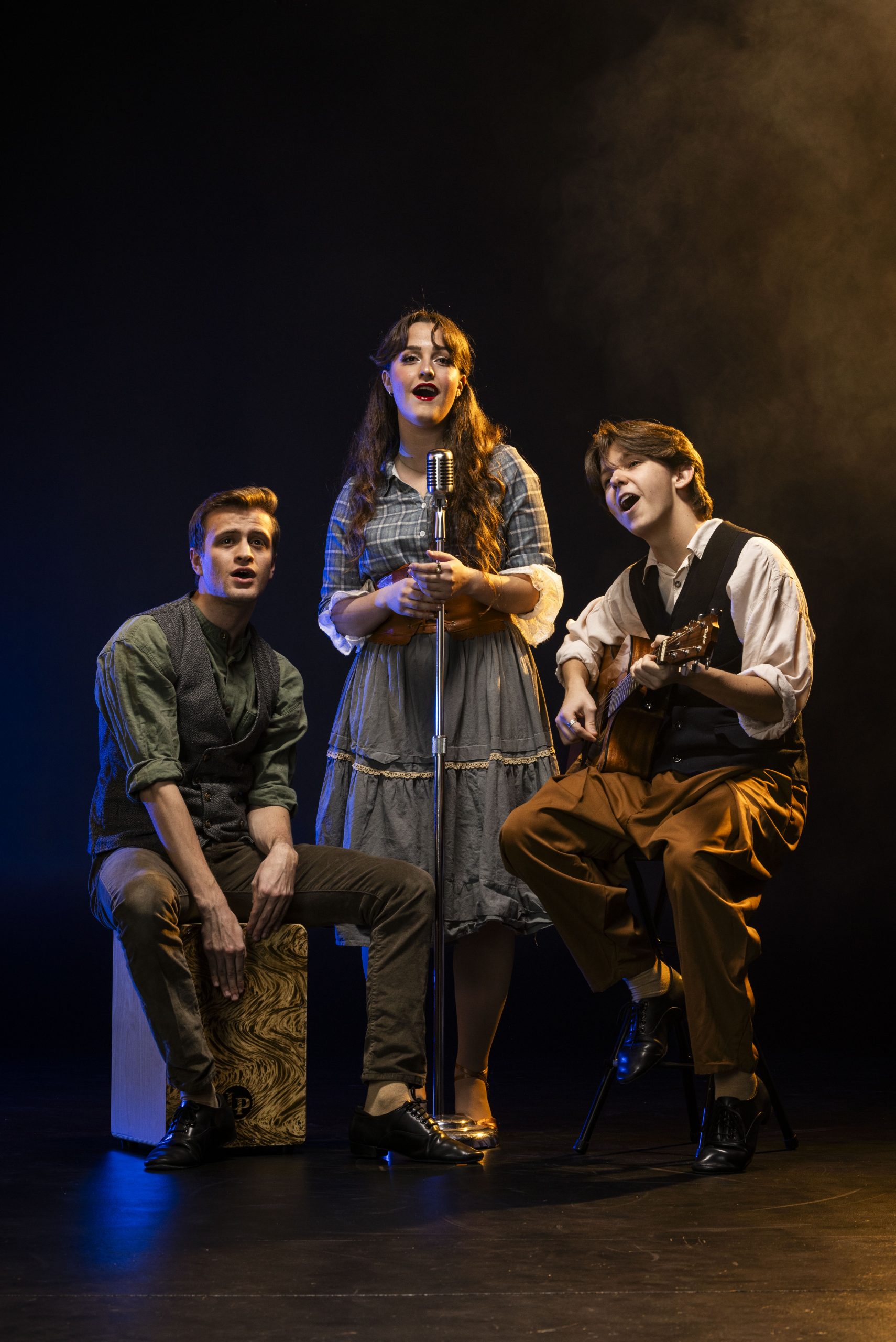 Three performers on stage singing together. One sits on a cajón, another plays guitar, and the third sings into a vintage-style microphone. They wear rustic costumes and are lit with dramatic stage lighting.