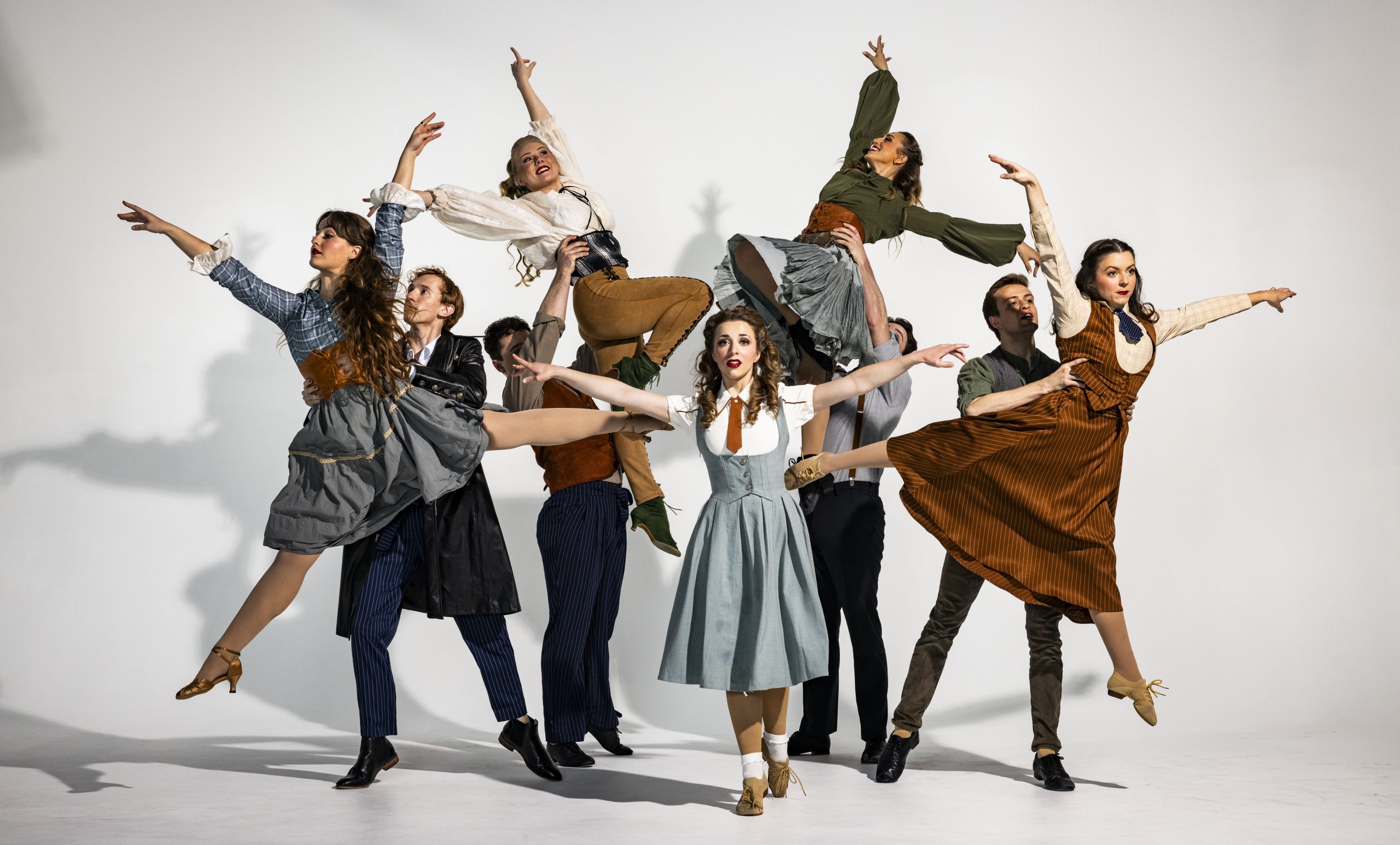 A group of dancers mid-performance, dressed in period-style costumes. Several women are lifted gracefully into the air while others strike expressive poses, creating a dynamic stage tableau.