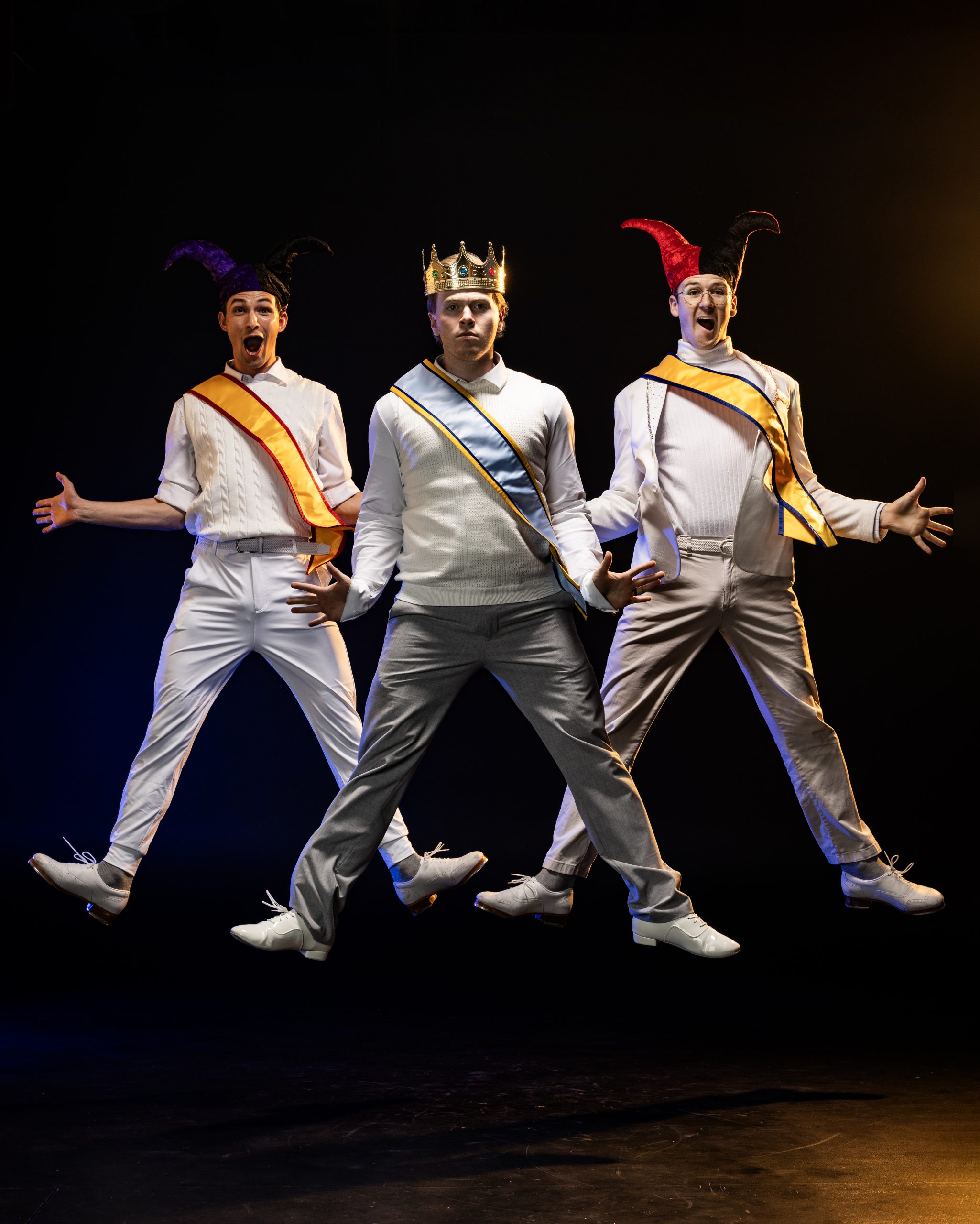 Three male performers mid-jump on stage. The center figure wears a crown and sash like a king, while the two on either side wear jester hats and sashes. All three have lively expressions and are lit by theatrical lighting.