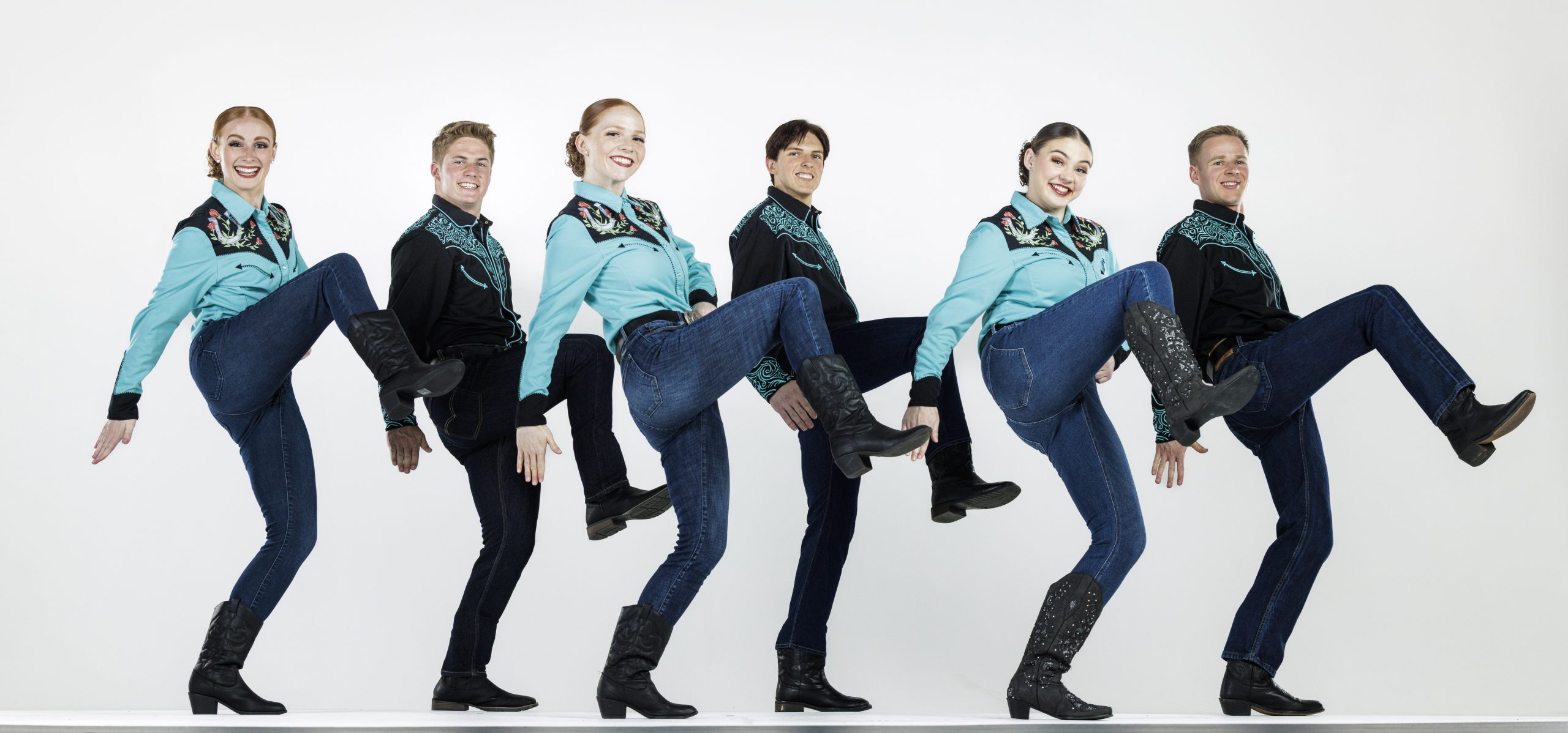 Six dancers in coordinated western outfits perform a high-kick routine. The women wear turquoise embroidered shirts, jeans, and black cowboy boots, while the men wear matching black embroidered shirts, jeans, and boots. All are smiling as they kick in unison against a plain white background.