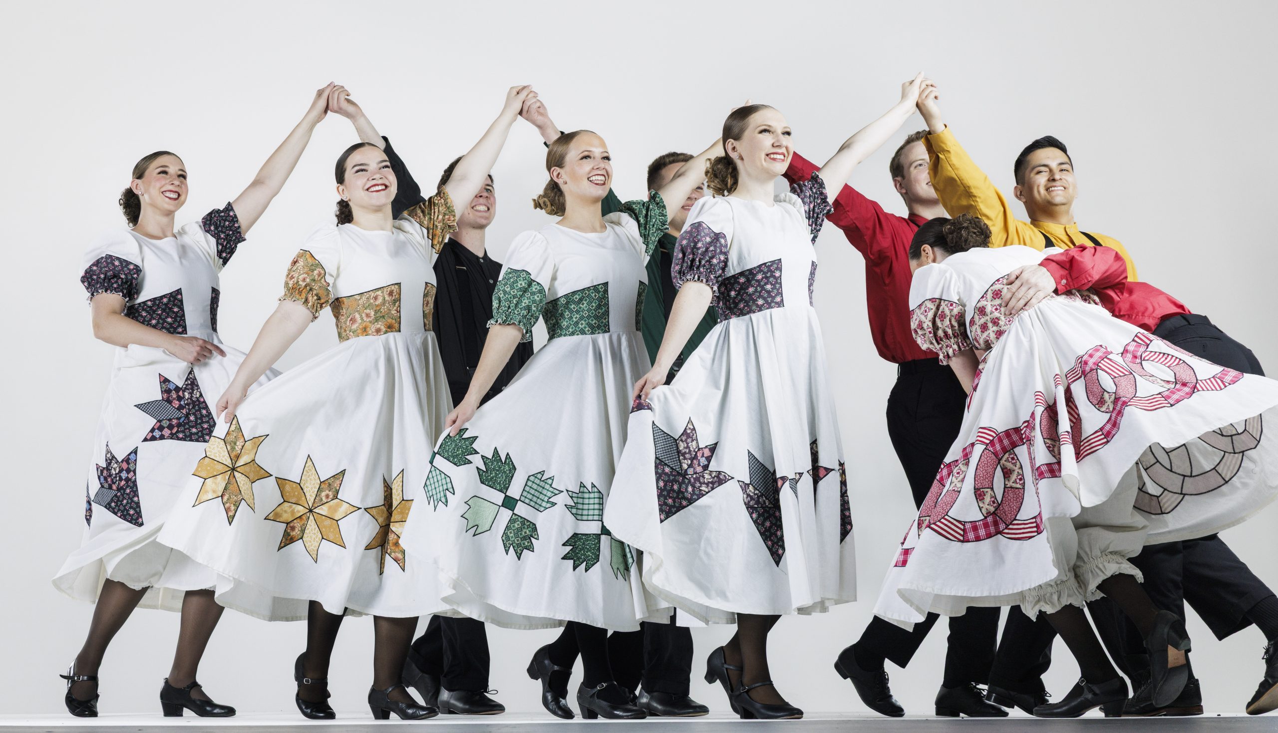 A group of dancers in pairs perform a folk dance, smiling as they hold hands in raised arches. The women wear white dresses with colorful quilt-patterned designs on the skirts and sleeves, while the men wear bright shirts in red, green, yellow, and black with suspenders. The dancers form a lively, flowing line against a plain white background.