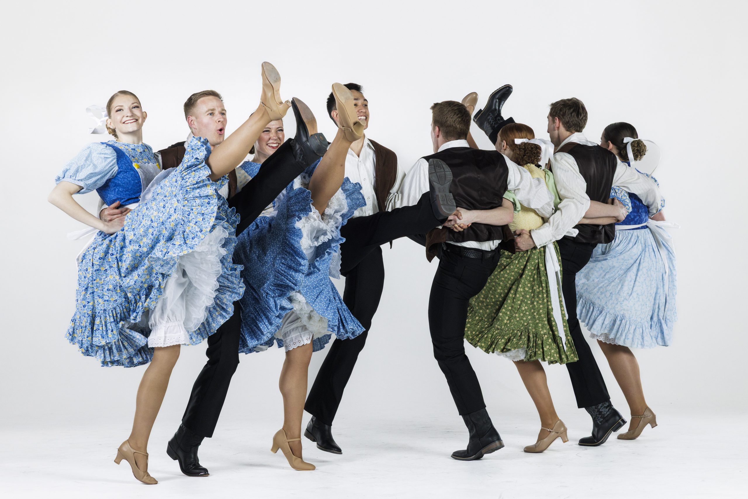 A line of folk dancers perform a high-energy routine. The women, wearing blue and green patterned dresses with ruffled skirts, kick their legs high in unison while the men, dressed in vests, white shirts, and black pants, hold them around the waist. The dancers are smiling and mid-motion against a plain white background.