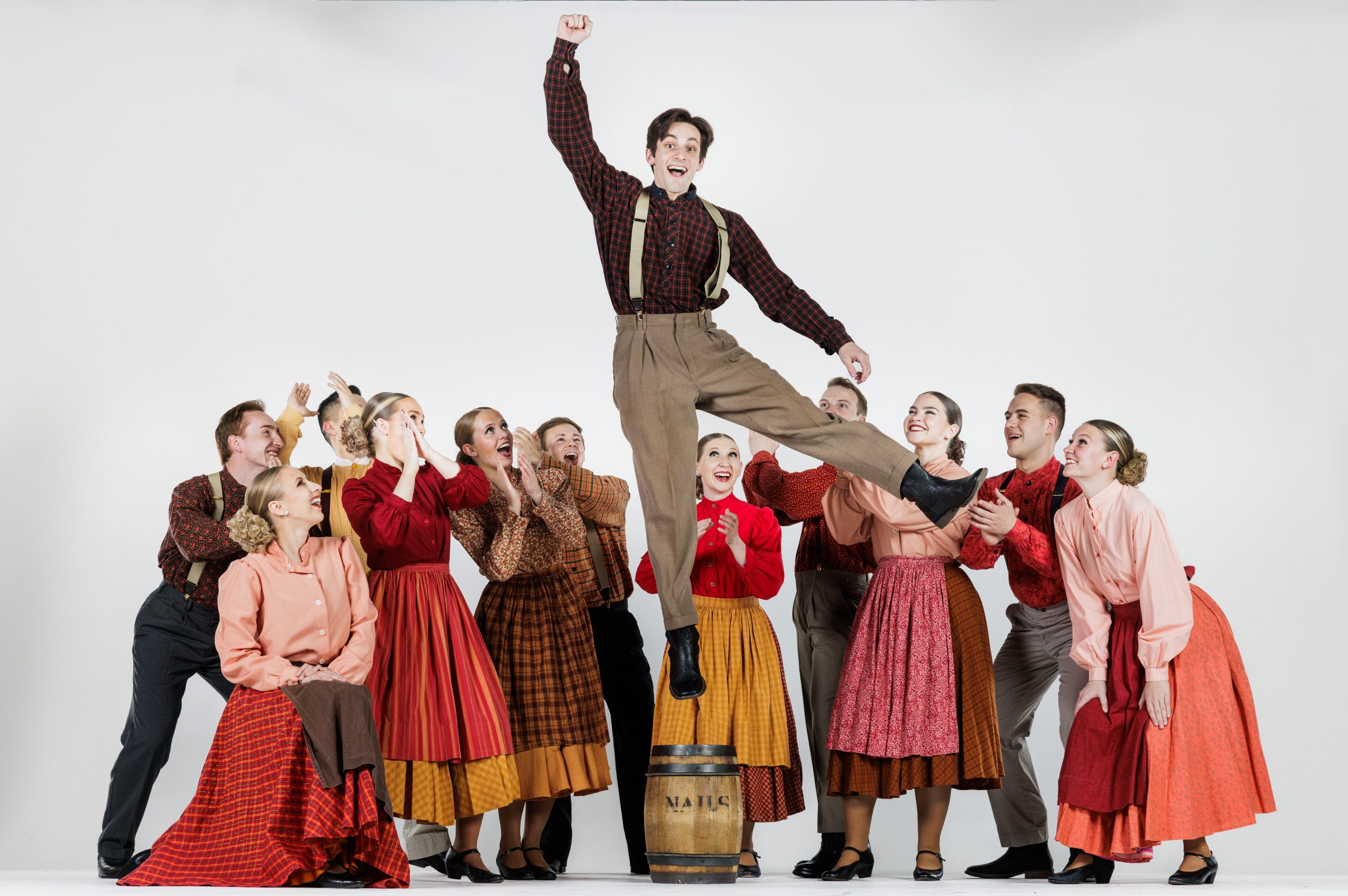 A group of folk dancers cheer as one male dancer leaps high into the air from a barrel, raising his fist triumphantly. The women wear long pioneer-style skirts and blouses in shades of red, orange, and brown, while the men wear coordinating shirts with suspenders. The group looks up in excitement against a plain white background