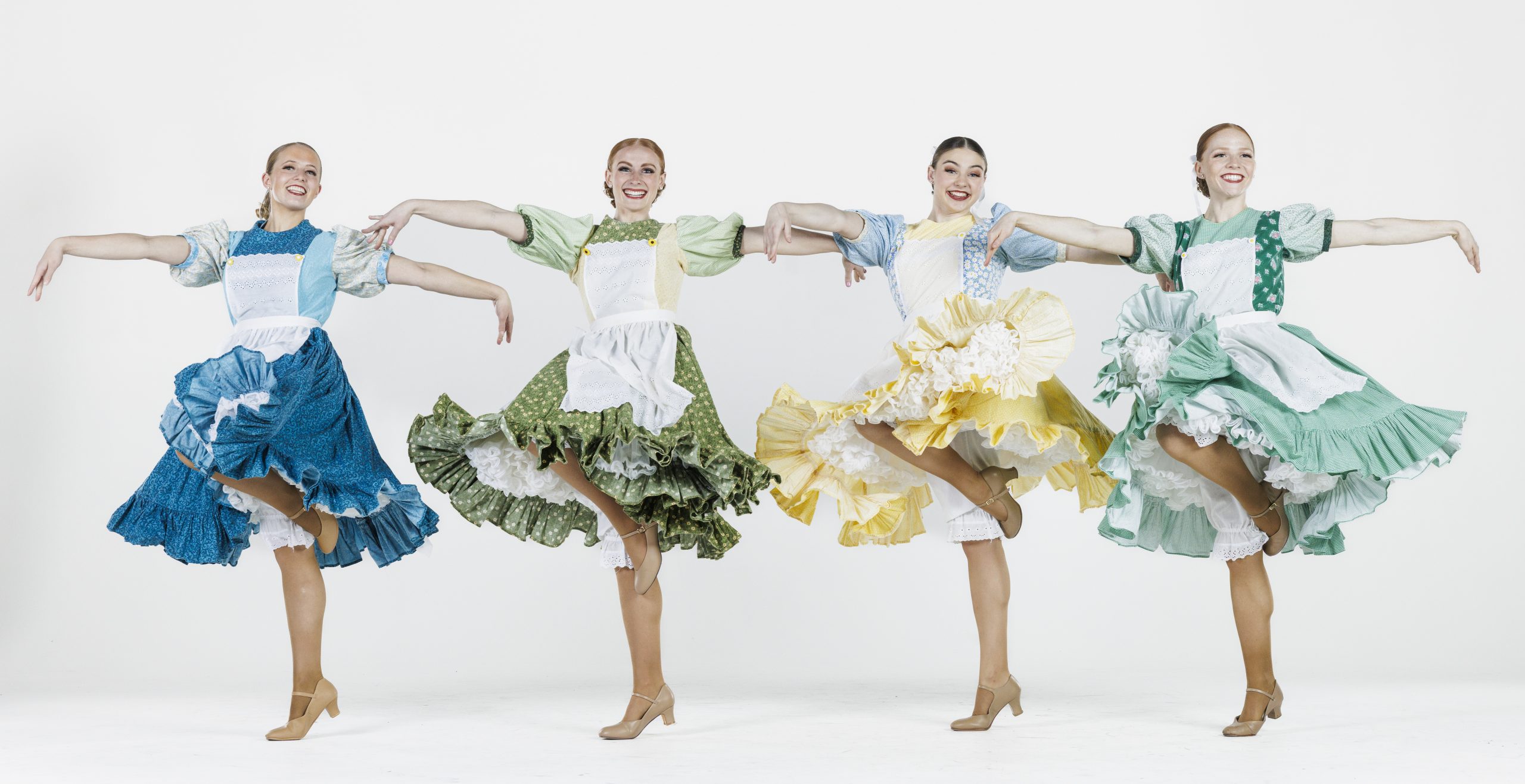 Four female dancers perform in colorful prairie-style dresses with aprons. Their skirts—blue, green, yellow, and teal—flare outward as they lift one leg and extend their arms gracefully. All smile brightly while dancing in unison against a plain white background.