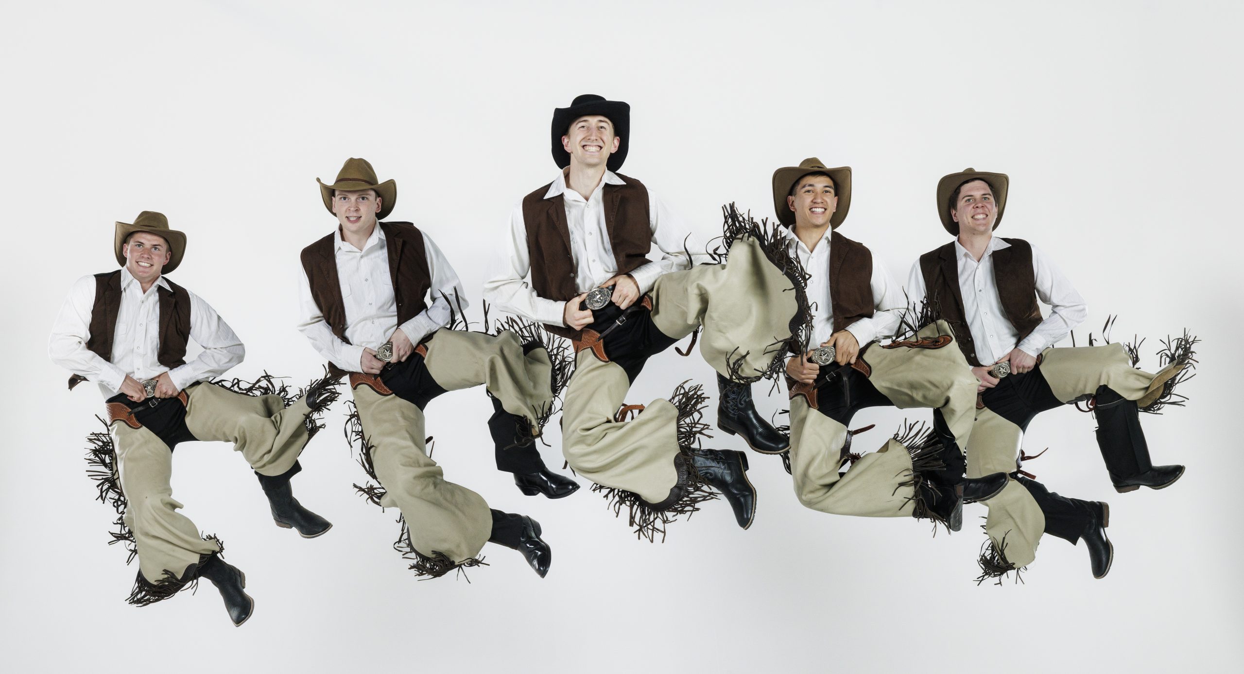 Five male dancers dressed as cowboys leap into the air in unison. They wear cowboy hats, white shirts, brown vests, tan chaps with fringe, and black boots, holding their belts with big smiles as they perform a high-energy western dance against a plain white background.