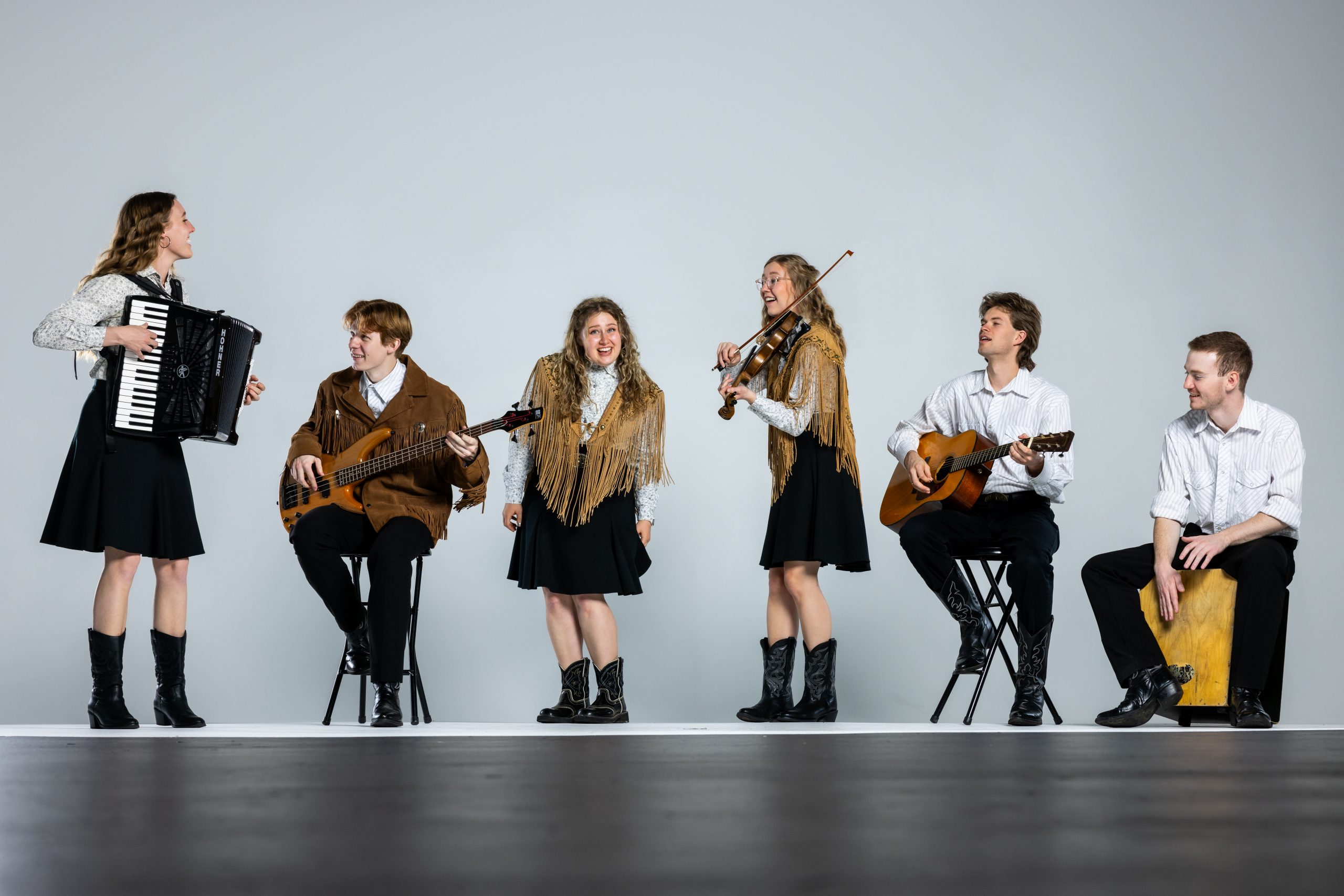 Mountain Strings players pose with their instruments (accordion, bass guitar, fiddle, acoustic guitar, and cajon) in traditional western dress against a white backdrop.