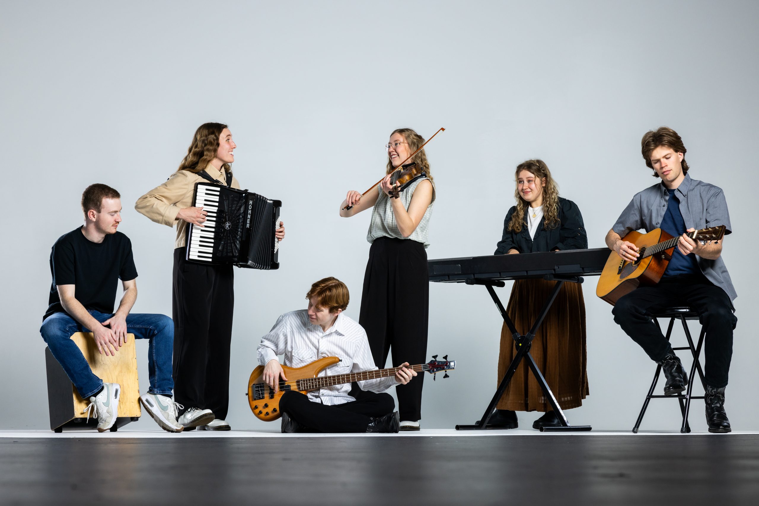 Mountain Strings members pose in casual dress against a white backdrop playing their instruments (cajon, accordion, bass guitar, fiddle, keyboard, and acoustic guitar).