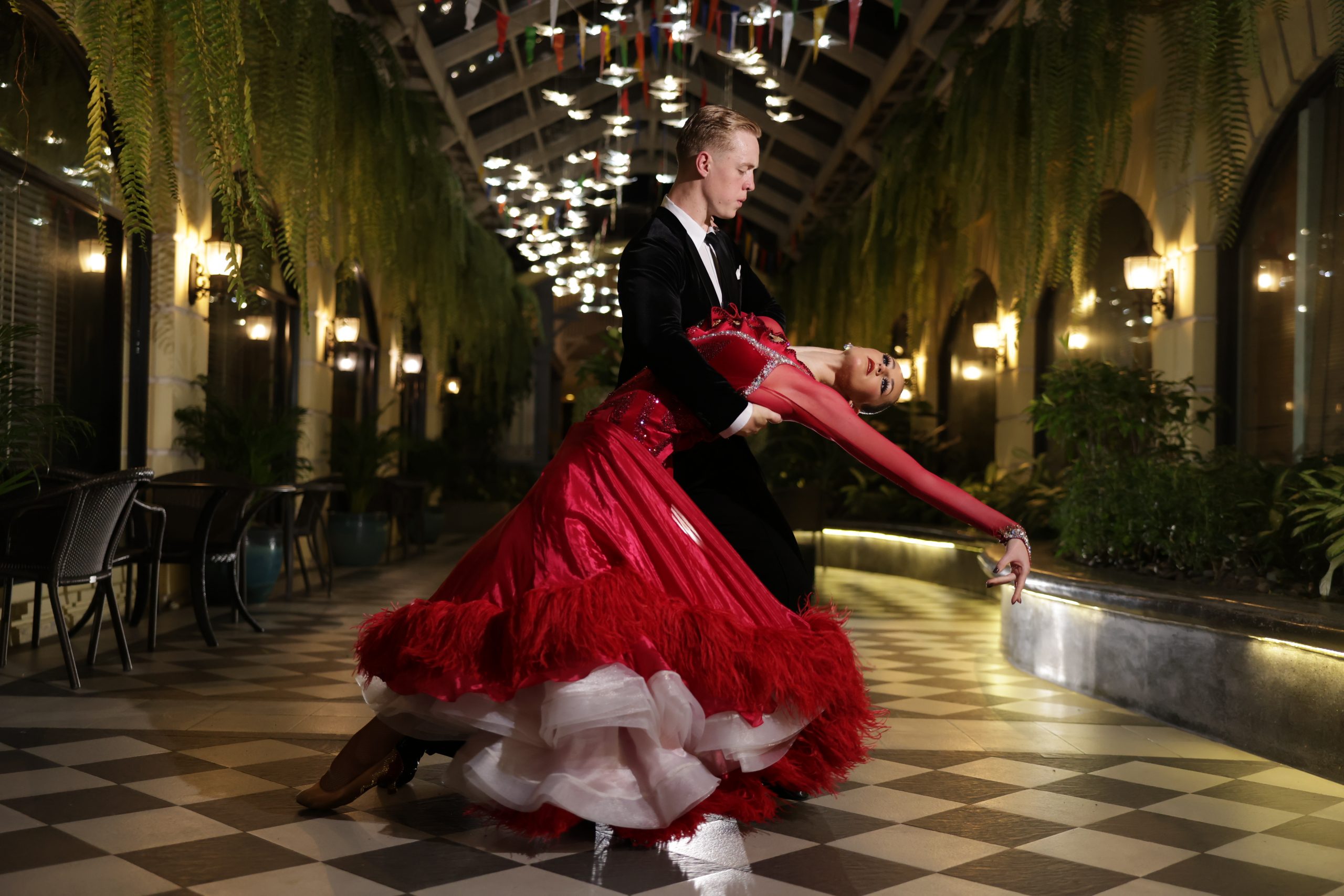 A ballroom dance couple performs a dramatic dip in an elegant hallway lined with plants and lights. The male dancer, dressed in a black tuxedo, supports his partner as she leans back gracefully in a flowing red gown with feathered trim and a full white underskirt. The checkered floor and glowing overhead lights add to the formal atmosphere.