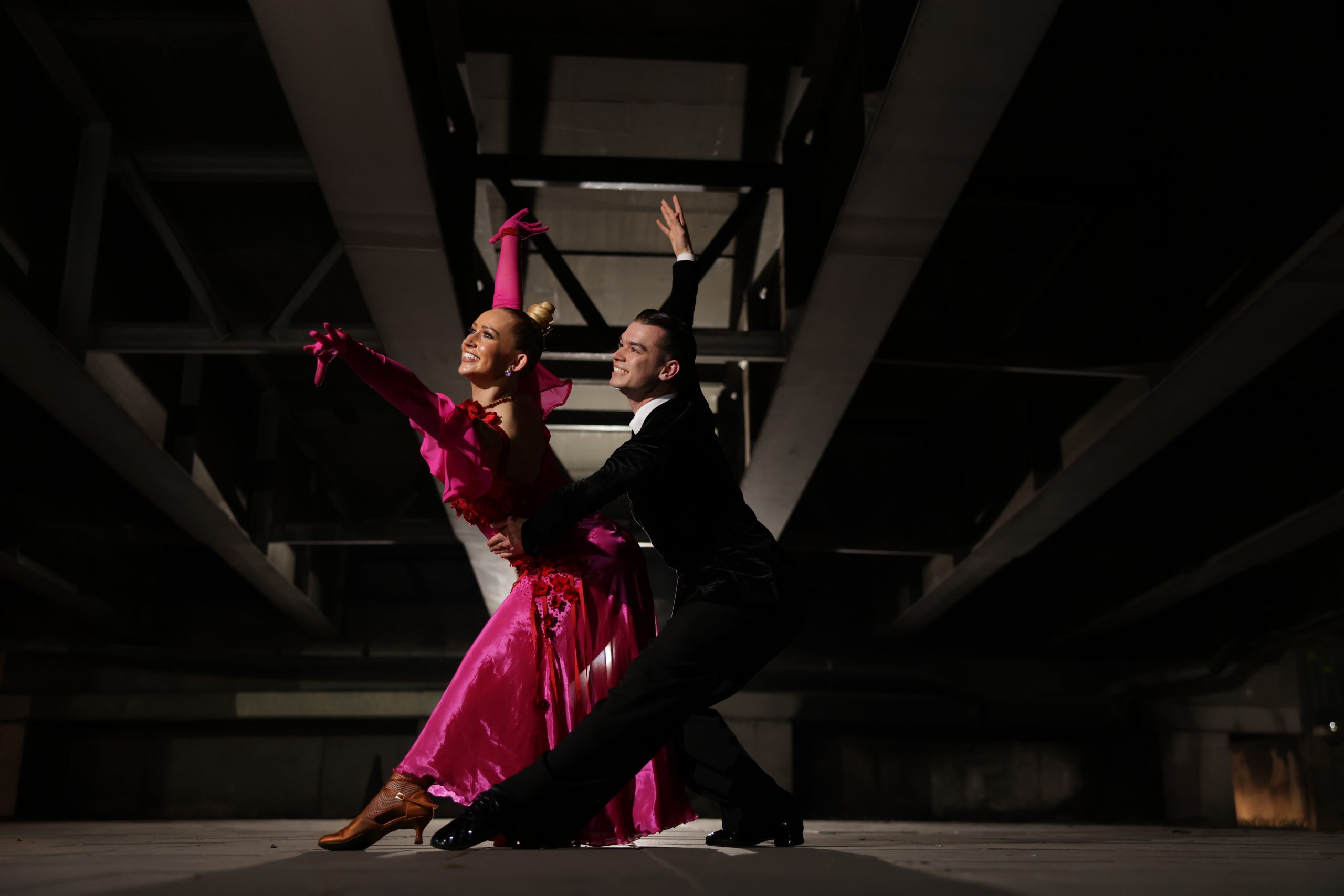 A ballroom dance couple performs dramatically beneath an industrial ceiling structure. The female dancer wears a flowing hot pink gown with long gloves, extending her arms upward as she smiles. The male dancer, in a black tuxedo, supports her in a deep lunge, also reaching one arm upward. Both dancers radiate energy and poise in the spotlight.