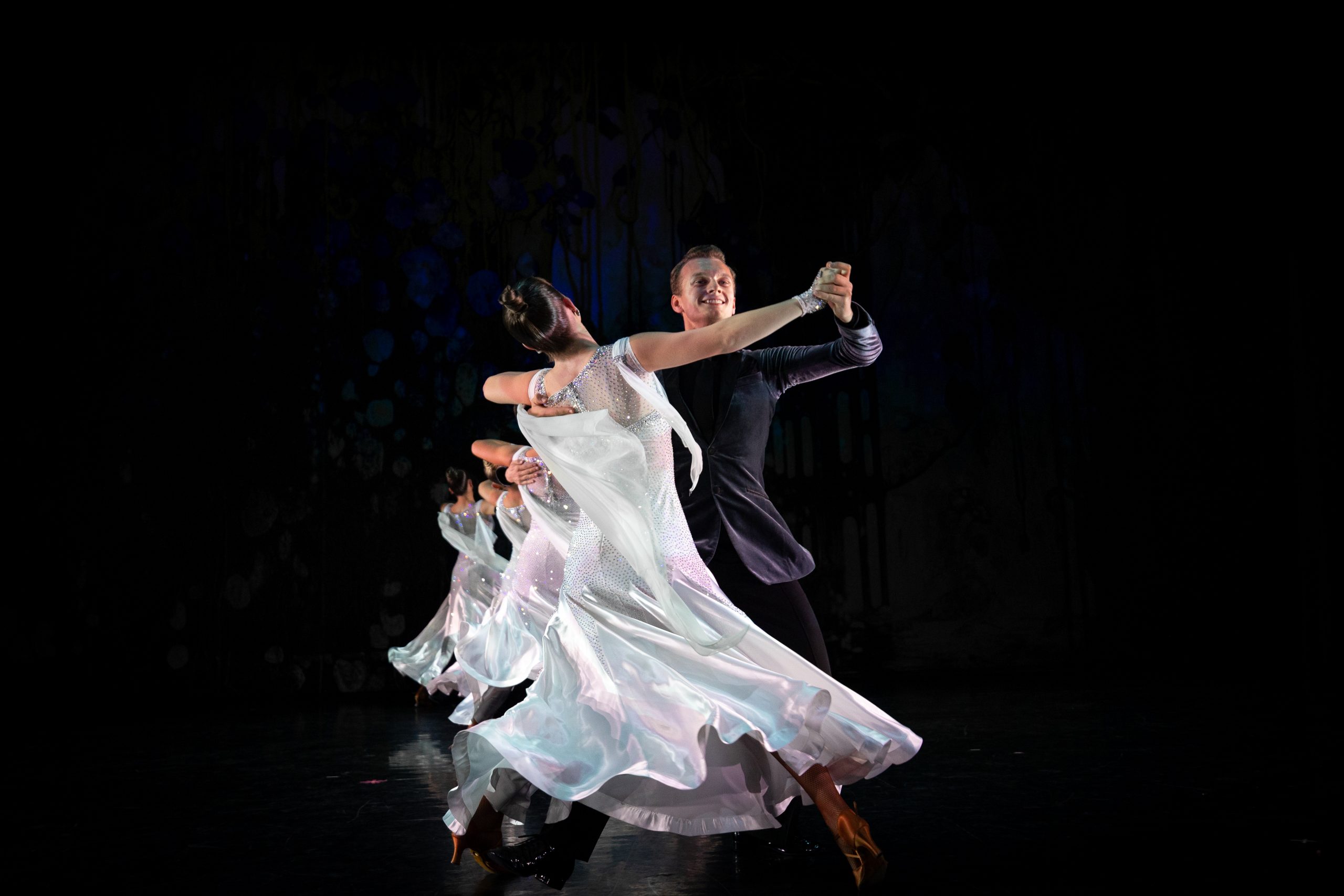 A ballroom dance couple performs a graceful waltz on stage. The female dancer wears a flowing white gown that swirls dramatically as she turns, while the male dancer, in a dark suit, holds her in a classic ballroom frame. Other couples in matching costumes dance in the background against a dark stage backdrop.