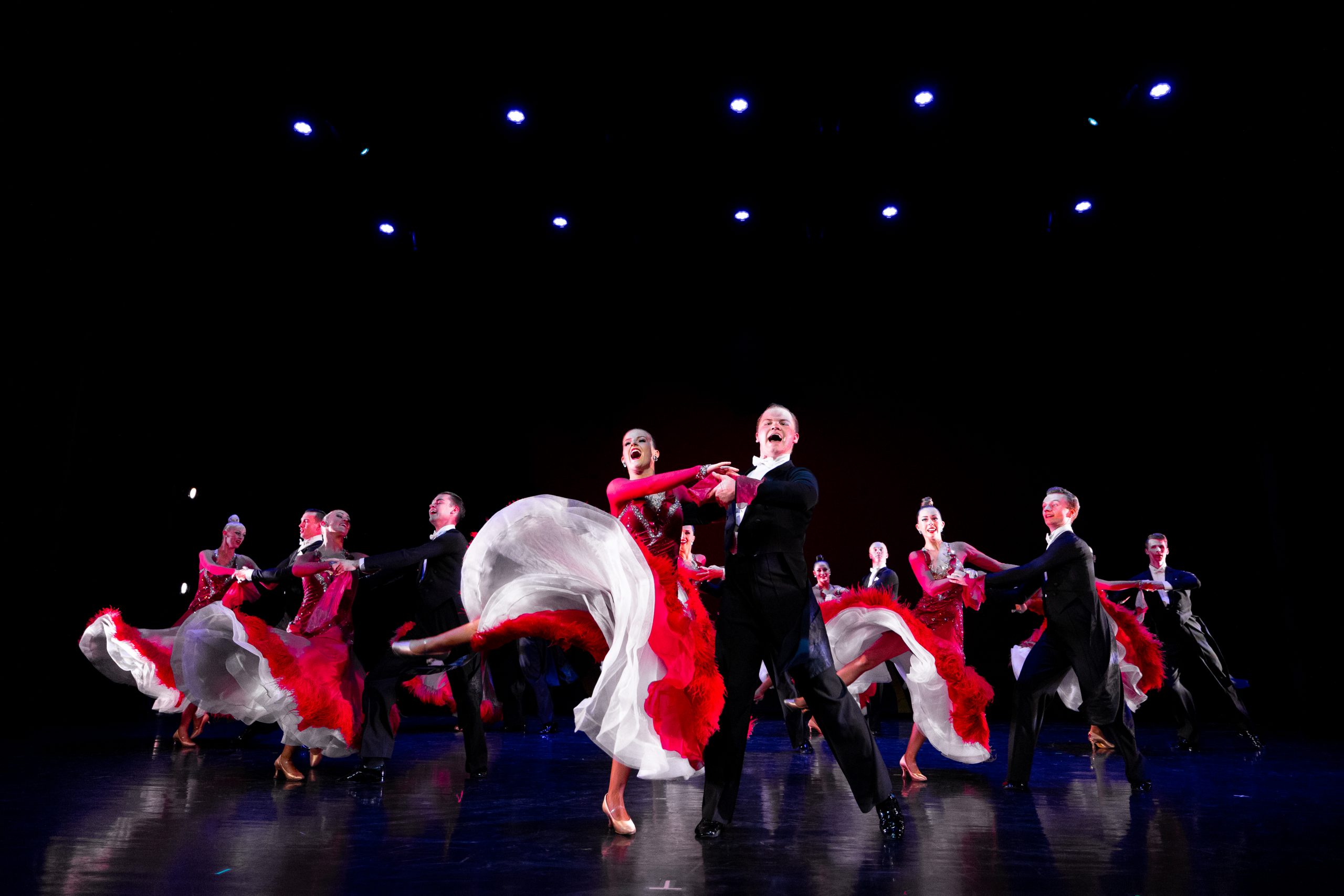 A group of ballroom dance couples perform energetically on stage. The women wear flowing red gowns with white underskirts that flare dramatically as they spin, while the men wear classic black tailcoats. The dancers move in unison under bright stage lights against a dark background, creating a dynamic, theatrical effect.