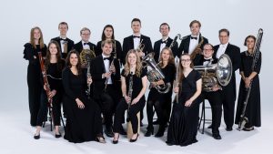 BYU Wind Symphony members gather around in concert black against a white studio back drop. The front row of musicians sit with instruments in their laps.
