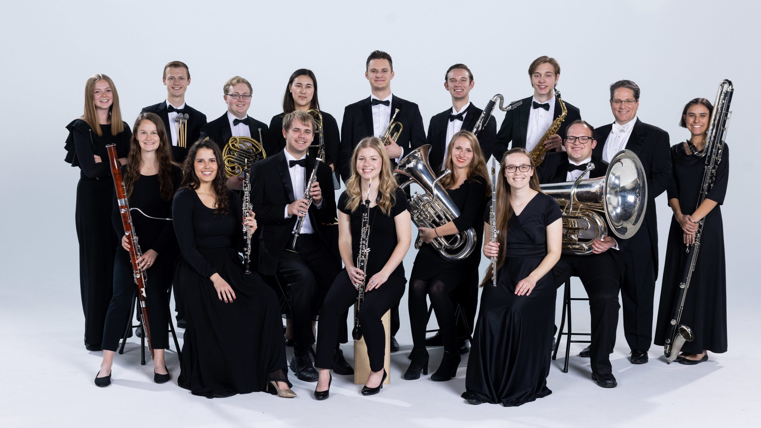 BYU Wind Symphony members gather around in concert black against a white studio back drop. The front row of musicians sit with instruments in their laps.